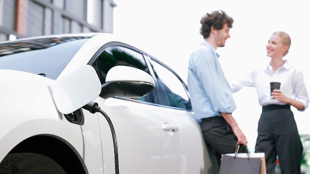 Uomo d'affari e donna d'affari progressisti che si appoggiano su un'auto elettrica collegata alla stazione di ricarica prima di guidare per il centro città Auto ricaricabile ecologica alimentata da energia pulita