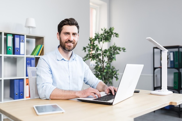 Uomo d'affari felice che lavora in un ufficio moderno e luminoso dietro l'uomo del computer portatile con la barba che sorride e che guarda le macchine fotografiche