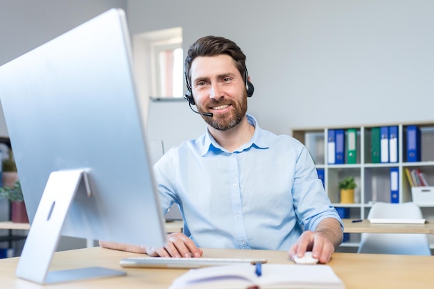 Uomo d'affari felice con auricolare per videochiamata che lavora in un ufficio moderno uomo sorridente che guarda il monitor del computer