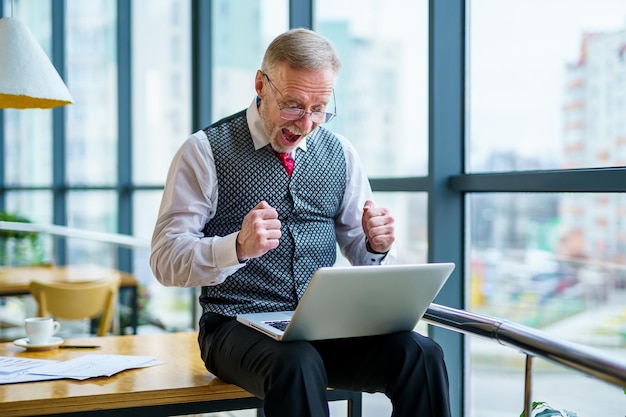 Uomo d'affari felice in giacca e cravatta guardando il computer portatile eccitato da buone notizie online fortunato vincitore di successo uomo seduto alla scrivania dell'ufficio alzando la mano nel gesto di sì che celebra il risultato di successo aziendale