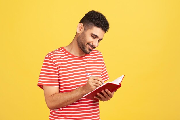 Uomo d'affari felice sorridente con la barba in maglietta a righe che scrive la lista delle cose da fare, prende appunti con la penna nel taccuino, controlla il programma. Studio indoor girato isolato su sfondo giallo