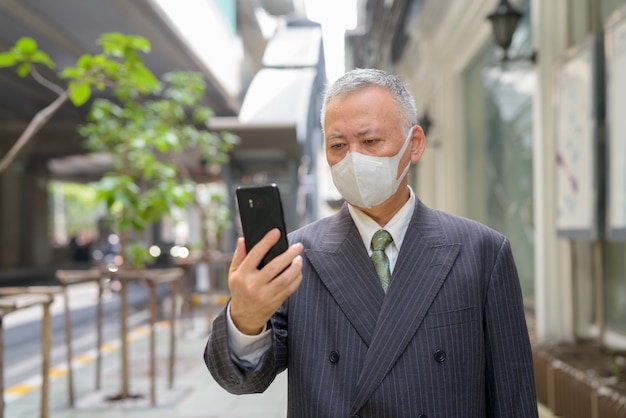 Uomo d'affari giapponese maturo con la maschera facendo uso del telefono nella città