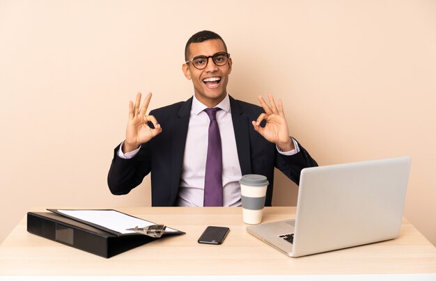 Uomo d'affari giovane nel suo ufficio con un computer portatile e altri documenti che mostrano un segno ok con le dita