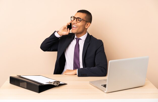 Uomo d'affari giovane nel suo ufficio con un computer portatile e altri documenti mantenendo una conversazione con il telefono cellulare