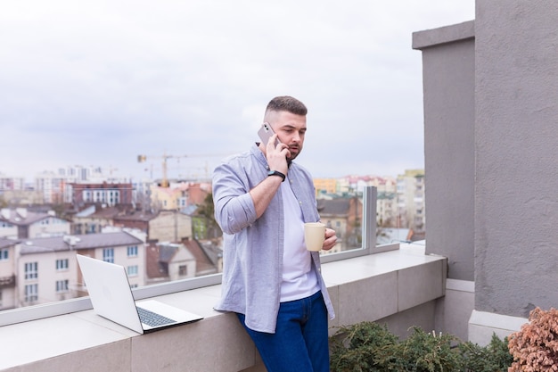 Uomo d'affari in abiti casual che si distende sulla terrazza con vista sulla città utilizza il telefono e utilizza un laptop mentre beve il caffè