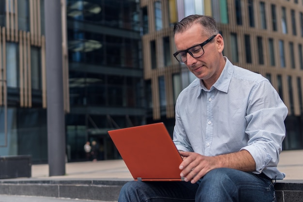 Uomo d'affari in abiti casual seduto sulle scale vicino a un edificio moderno e lavorando su laptop