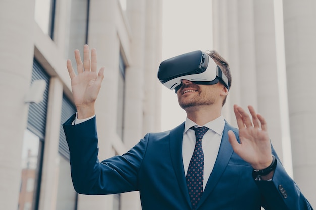 Uomo d'affari in occhiali per realtà virtuale che utilizza occhiali VR mentre si trova in una strada cittadina all'aperto, testando un metodo innovativo per il business, gesticolando mentre interagisce con l'interfaccia digitale