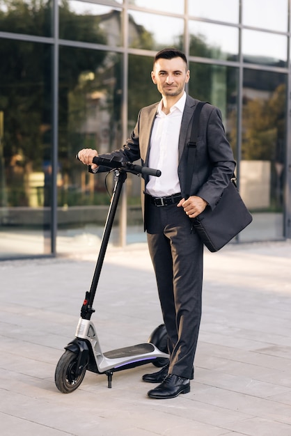 Uomo d'affari in piedi di fronte alla città che tiene in mano uno scooter elettrico e guarda la telecamera urbana moderna