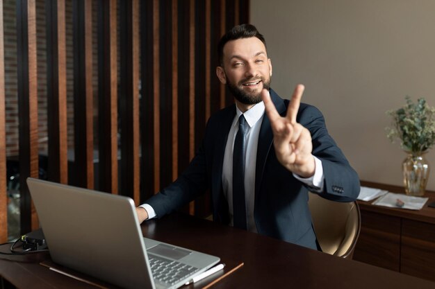 Uomo d'affari in un vestito seduto in ufficio davanti a un computer portatile che mostra un gesto di due dita