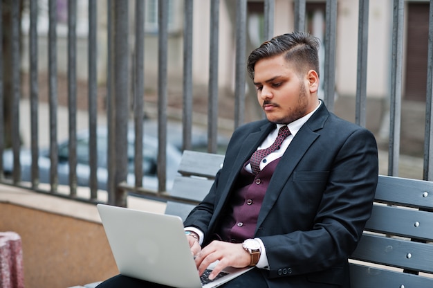 Uomo d'affari indiano alla moda nell'usura convenzionale che si siede sul banco con il computer portatile.