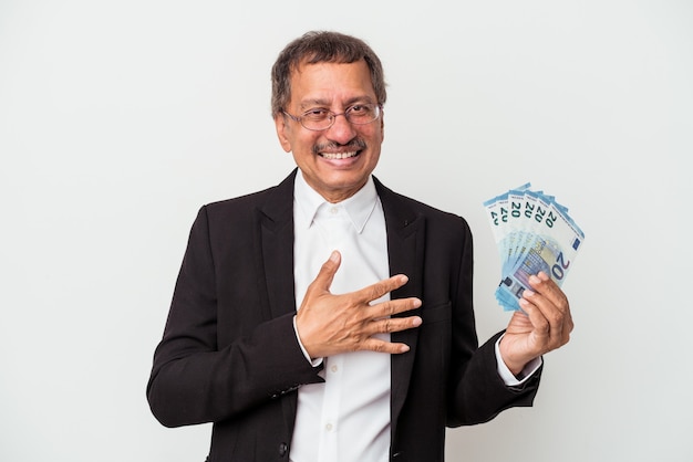Uomo d'affari indiano di mezza età che tiene banconote isolate su sfondo bianco ride forte tenendo la mano sul petto.
