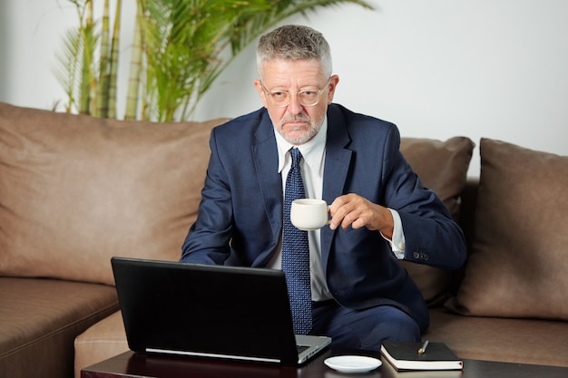 Uomo d'affari invecchiato infelice serio che beve tazza di espesso quando ha videochiamata mattutina con i colleghi delle filiali dell'azienda
