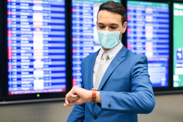 Uomo d'affari mascherato che controlla tempo nell'aeroporto, ritardo di volo durante il concetto di pandemia del coronavirus