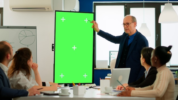 Uomo d'affari maturo che analizza i rapporti finanziari annuali in piedi nella sala conferenze che punta al monitor dello schermo verde. Leader che spiega la strategia del progetto utilizzando il display chroma key del mockup del pc nella sala riunioni