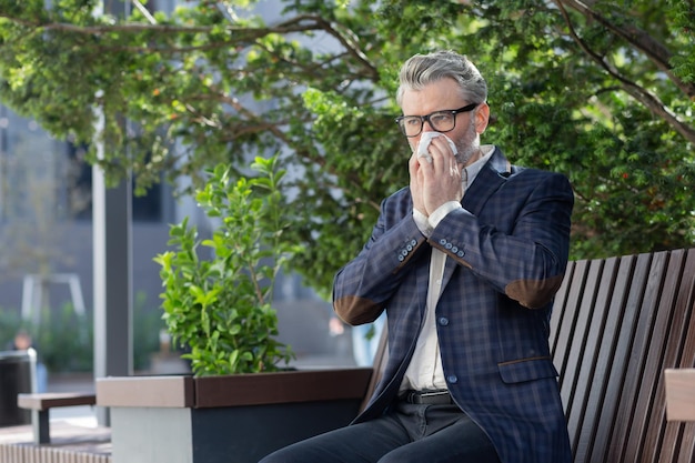 Uomo d'affari maturo che lavora all'aperto con il computer portatile uomo anziano che starnutisce dalle allergie seduto sul parco