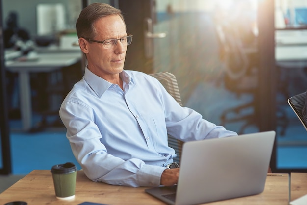 Uomo d'affari maturo concentrato in camicia blu che utilizza laptop mentre si lavora nell'ufficio moderno