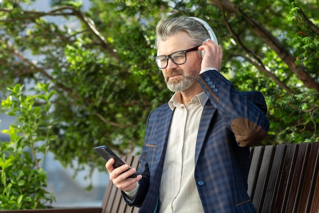 Uomo d'affari maturo e sicuro con le cuffie che usa lo smartphone in un parco urbano