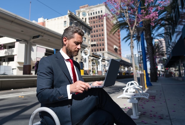 Uomo d'affari maturo in giacca e cravatta seduto su una panchina in città all'aperto che lavora online sull'educazione del laptop