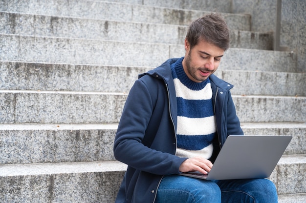 Uomo d'affari moderno dei pantaloni a vita bassa bei, usando computer portatile nella città, con l'espressione positiva.