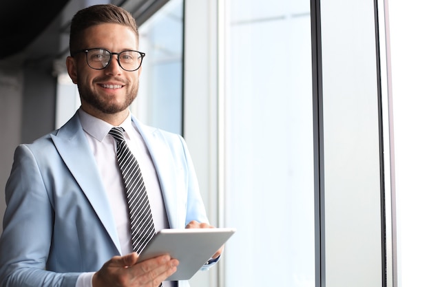 Uomo d'affari moderno in abiti da cerimonia che utilizza tablet digitale mentre si trova vicino alla finestra in ufficio.