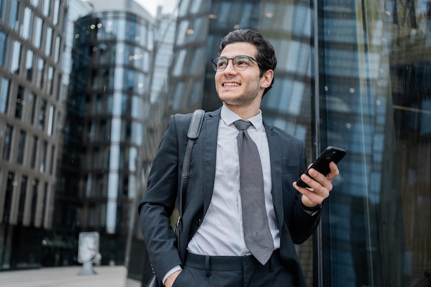 Uomo d'affari ottimista in abito che usa il telefono con edifici della città riflettenti sullo sfondo