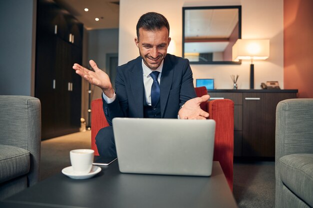 Uomo d'affari positivo in abito elegante seduto su una comoda poltrona con un laptop su un tavolino e gesticolando mentre sorride alla webcam