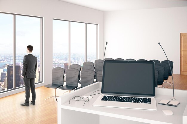 Uomo d'affari premuroso all'interno della sala conferenze con computer portatile vuoto su supporto con microfoni, occhiali e finestre con vista sulla città Mock up rendering 3D