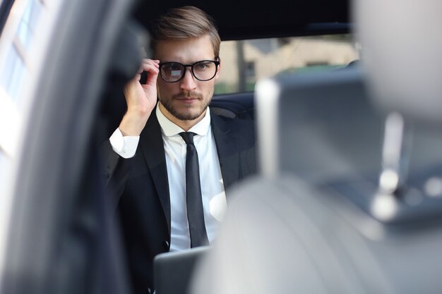 Uomo d'affari premuroso fiducioso che tiene la mano sugli occhiali mentre è seduto nell'auto di lusso.