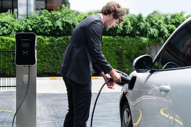 Uomo d'affari progressista che carica l'auto elettrica presso la stazione di ricarica per auto del parcheggio pubblico