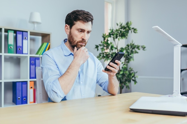 Uomo d'affari sconvolto e triste guardando l'uomo dello schermo del telefono che lavora in un ufficio luminoso