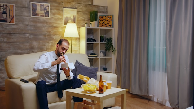 Uomo d'affari seduto sul divano a mangiare noodles da asporto dalla scatola dopo il lavoro nel suo appartamento a guardare la tv.