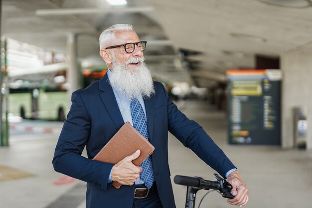 Uomo d'affari senior hipster che tiene in mano uno scooter elettrico mentre aspetta alla stazione degli autobus - Focus on face