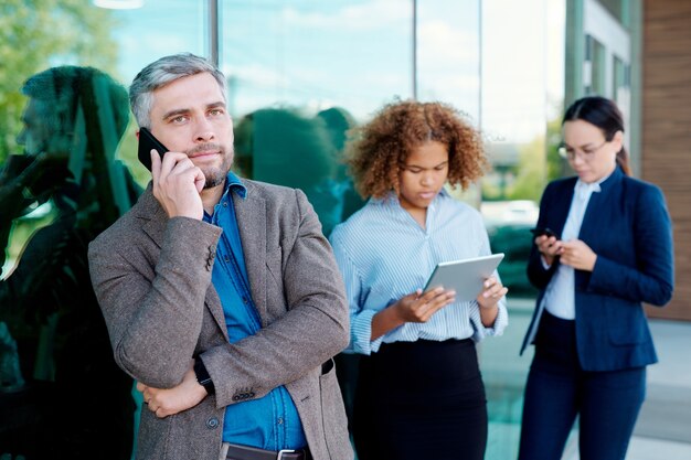 Uomo d'affari serio che parla sullo smartphone mentre fa una pausa dall'edificio per uffici sullo sfondo di giovani donne che utilizzano gadget