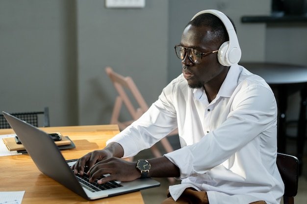 Uomo d'affari serio di etnia africana in camicia bianca e cuffie utilizzando laptop