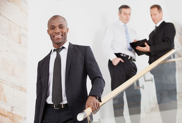Uomo d'affari sicuro. Allegro giovane uomo di colore in abiti da cerimonia che scende dalle scale e sorride mentre due persone parlano sullo sfondo