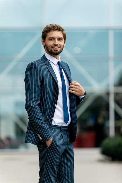 Uomo d'affari sicuro. Giovane sicuro in vestito pieno che regola la sua manica e che distoglie lo sguardo mentre stando all'aperto con paesaggio urbano nei precedenti