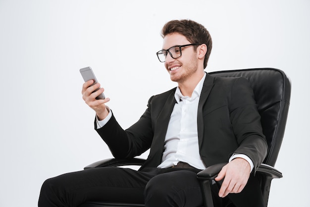 Uomo d'affari sorridente che usa il cellulare mentre è seduto su una sedia isolata sul muro bianco