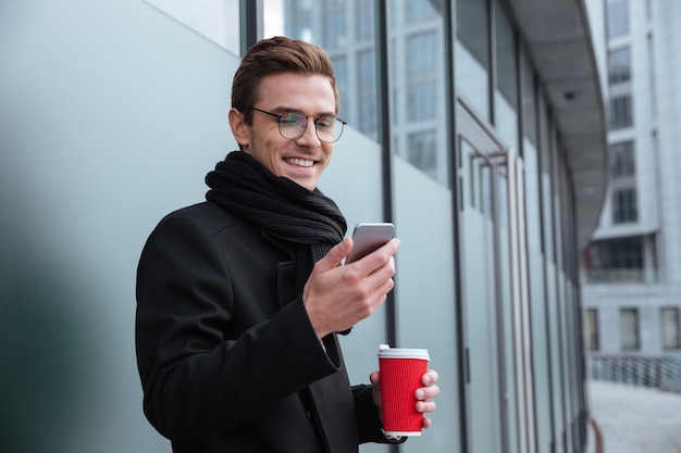 Uomo d'affari sorridente con gli occhiali e vestiti caldi con caffè e telefono all'aperto