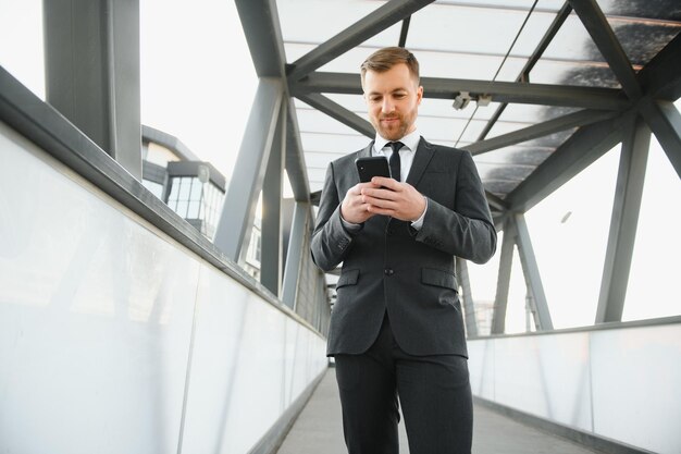 Uomo d'affari sorridente felice che indossa un abito nero e utilizza uno smartphone moderno vicino all'ufficio al mattino presto un datore di lavoro di successo per fare un affare mentre si trova vicino all'ufficio del grattacielo di notte la luce del chiarore