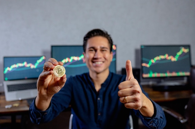 Uomo d'affari sorridente sullo sfondo che mostra una moneta di rappresentazione fisica bitcoin e il pollice in su. Buon investimento e concetto di trading.