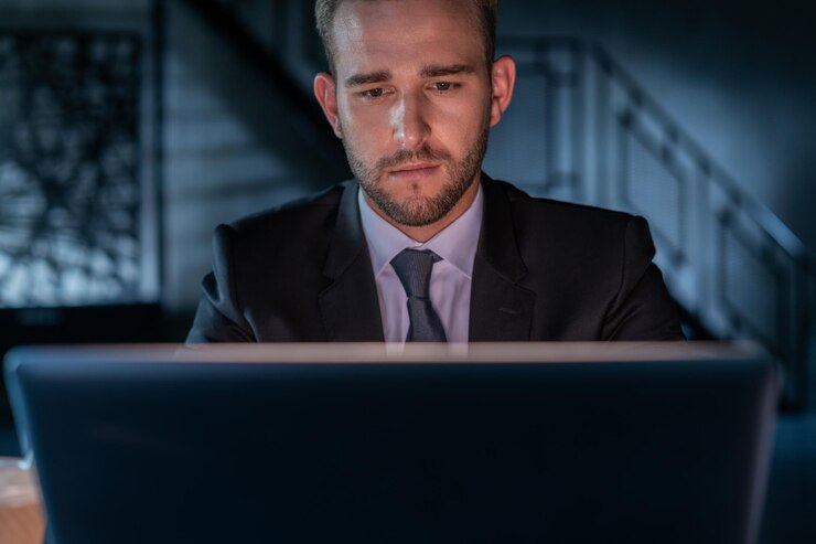 Uomo d'affari stanco che indossa un abito nero che lavora a tarda notte in ufficio Responsabile dell'ufficio che lavora con il computer portatile sfondo sfocato della stanza dell'ufficio buia Concetto di superlavoro