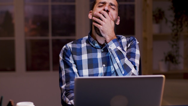 Uomo d'affari stanco che sbadiglia mentre lavora al suo laptop durante le ore notturne nel suo ufficio a casa.