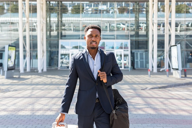 Uomo d'affari turistico afroamericano cammina edificio di trasporto pubblico con sfondo bagagli, vista posteriore posteriore L'uomo in abito formale viaggio in una città. Viaggiatore d'affari tirando la valigia nel terminal dell'aeroporto