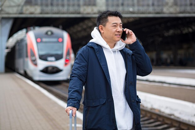 Uomo d'affari uomo asiatico alla stazione ferroviaria divertendosi a parlare al telefono un passeggero è arrivato in visita d'affari in una nuova città con una grande valigia di bagagli