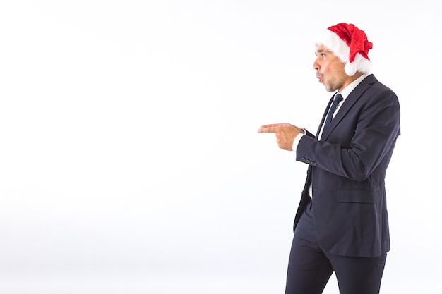 Uomo d'affari vestito con giacca, cravatta e cappello di Babbo Natale, sorpreso, indicando con le mani, su sfondo bianco. Concetto di celebrazione di Natale.