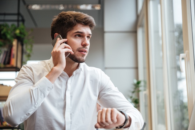 Uomo d'affari vestito con una camicia bianca seduto al bar e guarda da parte mentre parla al telefono