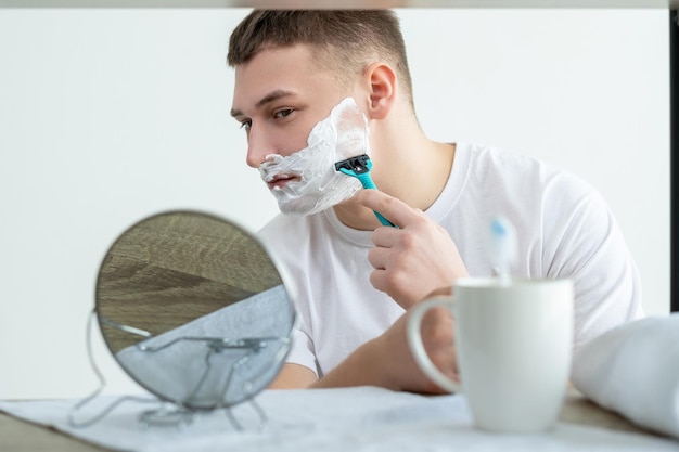 Uomo da barba Routine di cura della pelle Igiene del viso Ragazzo giovane e bello concentrato serio con schiuma sul viso che si guarda allo specchio usando il rasoio nel bagno bianco dopo la doccia