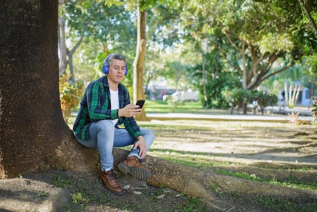 Uomo dai capelli grigi che ascolta musica con le cuffie e digita sul cellulare seduto in un parco accanto a un albero