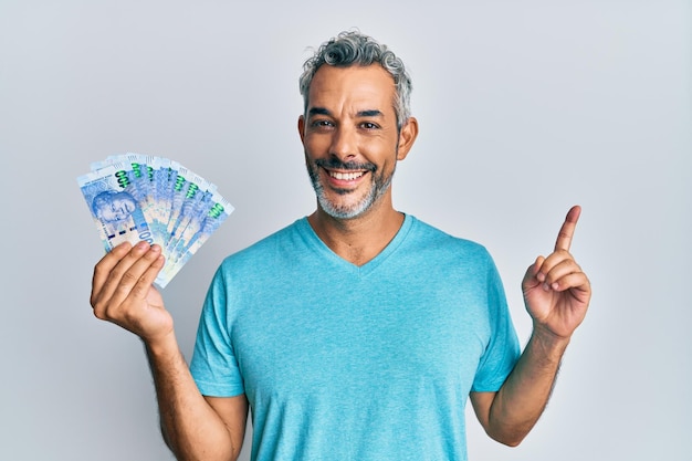 Uomo dai capelli grigi di mezza età che tiene banconote in rand sudafricani sorridendo felice indicando con la mano e il dito di lato