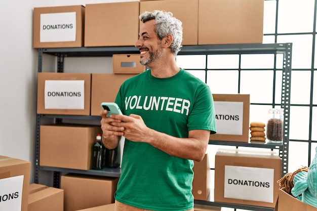 Uomo dai capelli grigi di mezza età volontario sorridente fiducioso utilizzando lo smartphone presso il centro di beneficenza